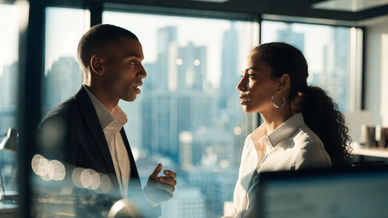 Color photo of a pest-control founder and a data analyst talking at a tall office window with bright city daylight behind them.
