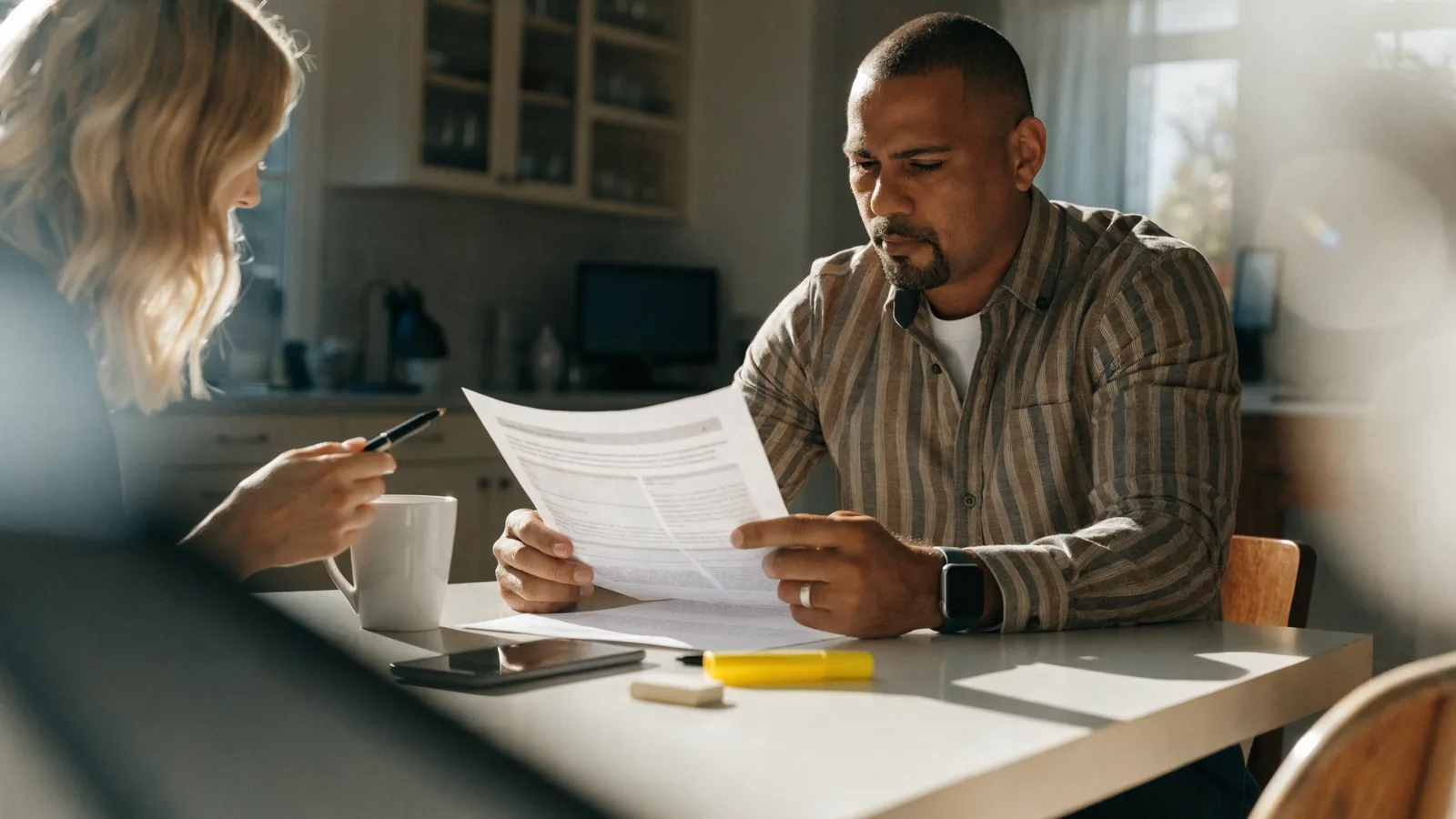 Color photo of a small pest-control business owner at a sunlit kitchen-table home office reviewing a printed quote with a customer across the table in bright morning light.