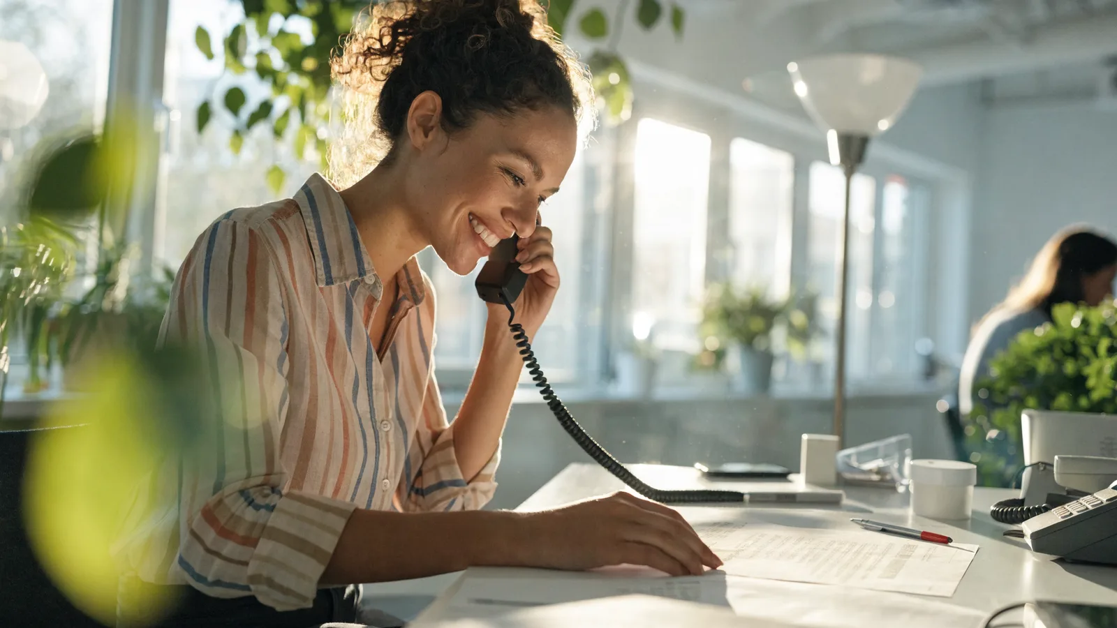 Photo of a pest control retention specialist on a phone call at her desk, reviewing a printed list of at-risk customer accounts while comparing Ardenus and RevHawk.
